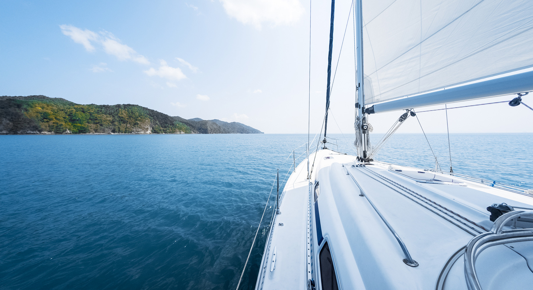 View from the deck of a sailing yacht heading toward open water, with calm blue seas, a distant green shoreline, and a clear sky with scattered clouds.
